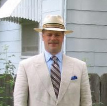 A middle aged man in a light tan suit wearing a straw hat, standing in a suburban backyard